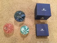 Three colorful blown glass paperweights (pink, blue, turquoise) shown alongside two blue Swarovski branded boxes on carpeted floor.