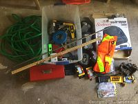 Wide view showing garden hose, plastic container with tools, red toolbox, safety vest, ruler and additional items on concrete floor.
