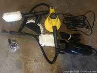 Yellow handheld steam cleaner with microfiber brush attachments and black hand vacuum with power cord on a concrete floor.