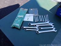 Photo showing 6 loose large combination wrenches arranged on a table alongside the 9 small wrenches in green plastic packaging.