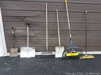 Front view of all six tools lined up against a brown wall on pavement: shovels, rakes, and broom visible.