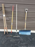 Photo of four long-handled gardening tools upright against a garage door: wooden post digger, yellow handled metal garden rake, white handled metal leaf rake, and blue blade metal shovel with wooden handle.