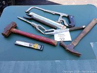 Photo of 6 miscellaneous tools including sledgehammer, hammer, wood chisel, coping saw, and U-shaped tool on green surface.