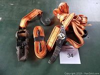 Three orange ratchet straps with metal hooks and ratchet mechanism, displayed on a dark green surface with lot number 34 in view.