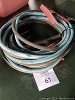 Coiled jumper cables with red and black clamps placed on a dark surface alongside a MaxSold lot tag number 61.
