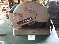 Front view of the Makita abrasive cutoff wheel mounted on a metal base, showing the protective wheel guard labeled with 'Makita'.