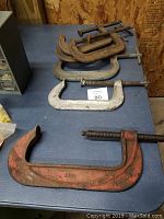 Six large metal C-clamps of various sizes displayed on a blue surface, showing a range of colors and signs of rust and wear.