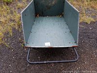 Front angled view of green wooden bed and black metal handle with two bicycle-style wheels on gravel ground
