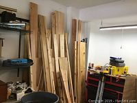 Photo of long vertical lumber pieces and plywood sheets leaning against garage wall with tools and equipment nearby
