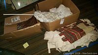 Boxes containing white linens with some soiling, large striped purse fabric on top, captured to show quantity and condition