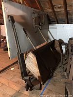 Photo of a large, hand-built wooden front loader with wooden handles and metal components, positioned in attic space with wooden floorboards and slanted roof.