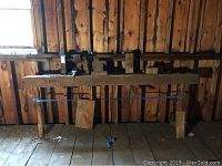 Full view of the large wooden log splitter mounted on legs with metal wedges and tools arranged on top, seen against wooden wall