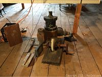 Collection of wooden and stone farm tools including anvil, mallet, grinding wheel, stones, and metal hand tools on wooden floor