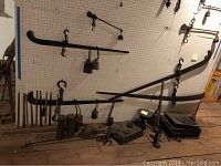 Wide side view showing large and medium black metal beam scales, various cast iron weights, hooks, and parts arranged on wooden floor and pegboard wall.
