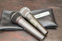 Two handheld dynamic microphones resting on a black pouch on a wood surface, one Shure SM58 and one Dixon DM-1, showing surface wear.