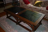 Front angle view of the wooden coffee table showing dark wood frame, two black rectangular tile inlays on the top, standing on a floral carpet near a sofa.