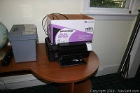 HP PhotoSmart printer sitting on wooden desk with a box of paper and plastic bin next to it.