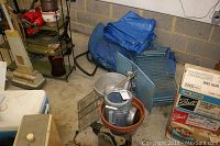 Wide view showing blue tarps, watering can, metal canteens, wire racks, flower pot, and boxes on concrete floor against cinder block wall