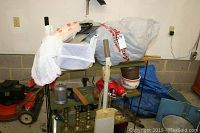 Metal shelf holding white small artificial Christmas tree, metal kitchen containers, brass candle holders, flower pots, Kenmore floor machine, gloves, and other miscellaneous items in a basement corner.