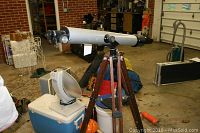 Telescope mounted on wooden tripod, displayed in garage with other items in background.