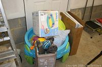Wide shot of pack and play with boxed potty step stool on top and beach toys inside, with folded shelving unit and cardboard boxes behind.