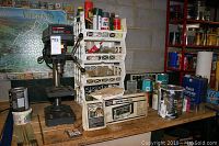 Wide view of workbench showing drill press on left, assorted paint cans, spray cans in white plastic rack, and ignition analyzer box at center.