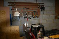 Photo of toolbox area with various bar clamps, hand tools hanging on pegboard wall, red hydraulic floor jack, and black bench vise on table.
