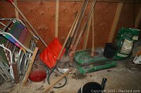 Wide view showing red plastic wheelbarrow, gardening tools with wooden handles, bags of garden soil, and folding lawn chairs against the wooden shed wall with floor dirt and leaves.