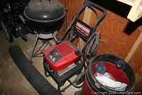 Craftsman pressure washer with red engine cover, black frame, and attached hoses near a black grill and red bucket.