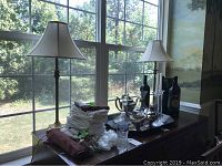 Two brass colored metal table lamps on wooden cabinet in front of window with outdoor greenery background. Lamps have cream fabric shades and round bases.