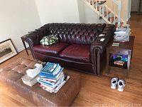 Photo showing burgundy leather chesterfield sofa with end table positioned beside it on wooden floor. End table is medium brown wood, rectangular with one drawer.