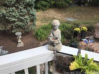 12-inch tall seated child concrete statue sitting on a porch ledge with outdoor plants in background.