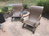 Two brown stained wooden Plantation rocking chairs showing front and side views set on outdoor brick patio with grass and plants in background.
