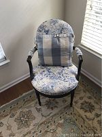 Front view of wooden upholstered armchair with toile fabric and blue checkered pillow on seat, showing padded arms and backrest.