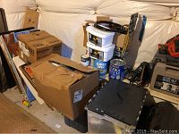 View of stacked cardboard boxes, used paint cans including Super Spec and Extra Deluxe interior paint, a Wagner power painter, and plastic storage tubs containing decorating supplies.