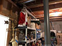 Overall view showing mechanical Santa Claus on top shelf, brass candle stakes and weather vane to the left, shelving with various holiday decorations.