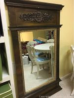 Full view of the ornate carved dark wood framed beveled mirror showing its rectangular shape and size in a room with chairs and table.