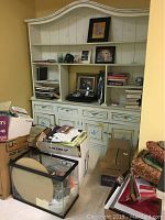 Photo showing wooden painted cabinet with decorative molding filled with books and framed photos, boxes of books, a fish tank and other items on floor.