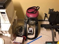 Full view of iGallop machine showing black frame with pink padded seat, fitness accessories including resistance bands, weight plates, boxed boxing gym accessory, and Pilates DVDs next to it.