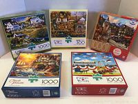 Photo shows all five jigsaw puzzle boxes arranged in two rows. Front row includes Chuck Pinson and Charles Wysocki puzzles, with Milton Bradley German Market Town and Charles Wysocki Olde Buck's County puzzles behind.