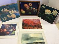 Five artwork prints arranged on white surface. Three prints shown: two on wood blocks with white flowers on dark backgrounds, one wooden framed still life with apples in basket, and two prints on paper with abstract and landscape scenes.