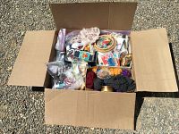 Top-down shot of a cardboard box filled with sewing and crafting supplies, showing yarn, hoops, trims, ribbons, and packaged needles.