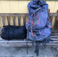 Photo showing large grey backpack with red straps and tightly rolled black sleeping bag beside it on a wooden bench.