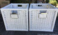 Two white wicker rectangular baskets with woven texture, metal label holders front, and cut-out handles on sides, shown outdoors on wooden surface.