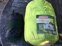Photo of two packed items on a wooden surface: a bright yellow/green Coleman sleeping bag cover with label and a smaller black and green pouch containing fleece blanket.