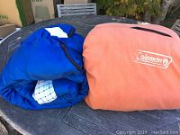 Photo of two sleeping bags rolled and placed side by side on a table, showing the Coleman logo on the orange bag and the blue checkered pattern of the other bag inner lining