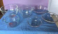 Photo showing six clear glass kitchen bowls on blue fabric - includes mixing bowls and measuring vessels of various sizes and brands.