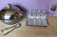 Photo showing all barware items: aluminum ice bucket with lid and wooden knob, eight small clear stemmed glasses on a silver and black serving tray, long stemmed brown tinted glass, and ice tongs.