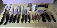 Overview of all knives and kitchen tools arranged on wooden surface including 11 knives, pizza cutter, cheese slicer, and two knife sharpeners.