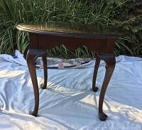 Brown wooden side table with curved legs on a white sheet outdoors, showing polished top and overall shape.
